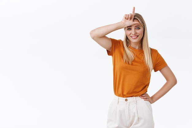 Snobistische knappe achteloze stijlvolle moderne blonde vrouw in oranje t-shirt met tatoeage houd hand op taille toon verliezer L woord op voorhoofd en lachend met minachting spottende vriend voor het verliezen