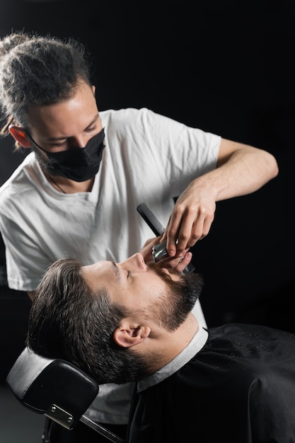 Snor scheren in kapperszaak. Kapper met dreadlocks in zwart medisch masker trim baard van knappe man bij quarantaine coronavirus covid-19.