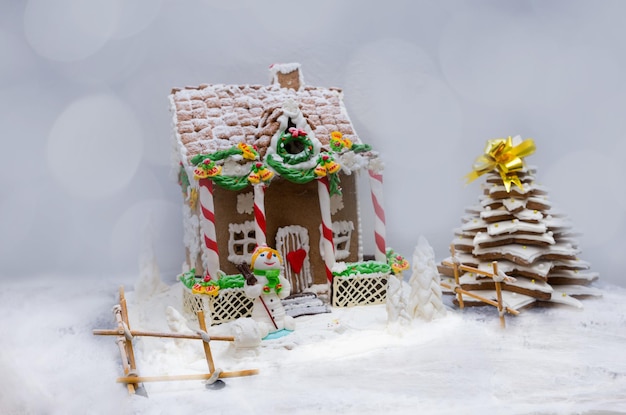 Snow-covered homemade gingerbread house, gingerbread Christmas tree and a sugar mastic snowman on defocused background. Mockup for seasonal offers and holiday post card