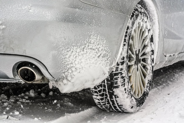 Snow covered rear wheel tire of silver car standing on winter ground, closeup detail