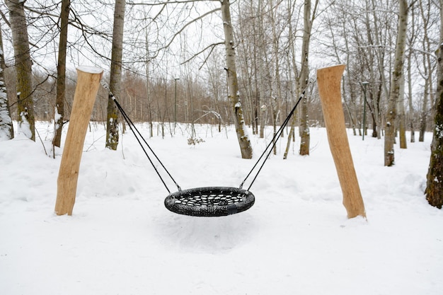A snow-covered swing in the old style in the form of a lounger  suspended from large logs with halpe ropes. Winter forest landscape.