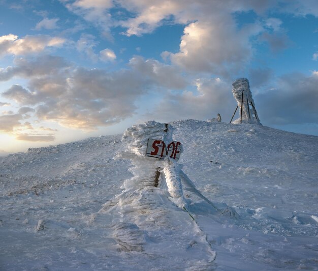 吹雪の前の最後の夕方の日光で冬の山の頂上は雪で覆われていますポインター上の風の強い霜と危険な崖に関する一時停止の標識