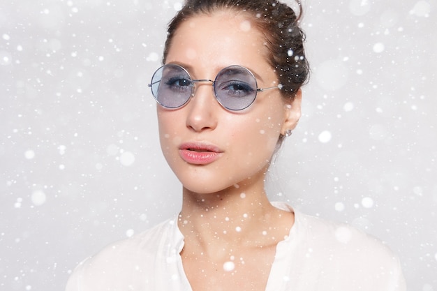 Snow, winter, christmas, people, beauty concept- Pretty young girl in white blank t-shirt, wearing glasses, empty wall, studio portrait. Smiling brunette woman in eyeglasses over snow background