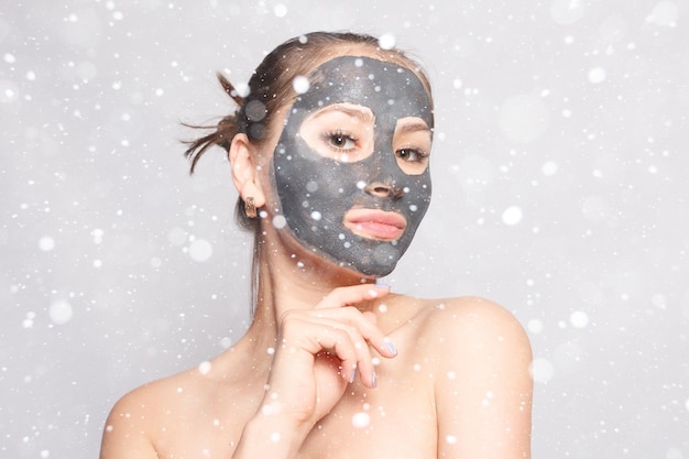 Neve, inverno, natale, persone, concetto di bellezza: maschera viso donna. ritratto di una bella ragazza che rimuove la maschera peeling nera cosmetica dalla pelle del viso su sfondo di neve