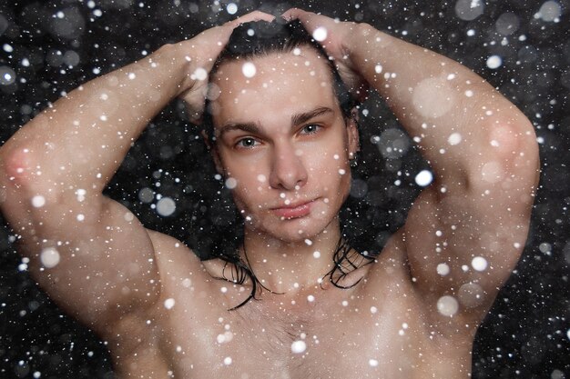 雪、冬、クリスマス、人々、スキンケア、美容のコンセプト-黒い雪の背景に長い黒髪の濡れた若い男。剃った胸を持つ肖像画の男性。男性のスキンケア。