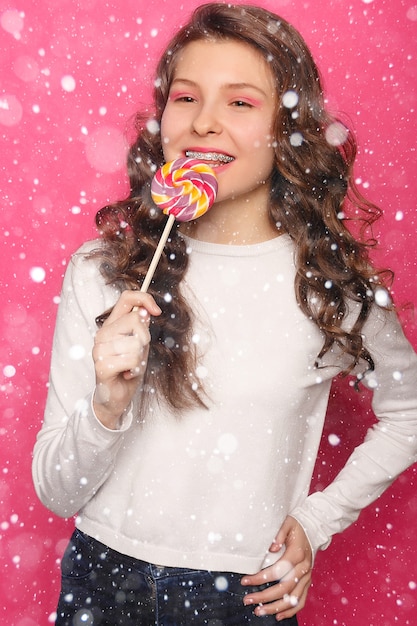 雪、冬、クリスマス、歯、感情、健康、人々、歯科医、ライフスタイルのコンセプト-ロリポップを保持している歯列矯正器を持つ女性。雪の背景の上の明確なブラケットと健康的な笑顔の女性