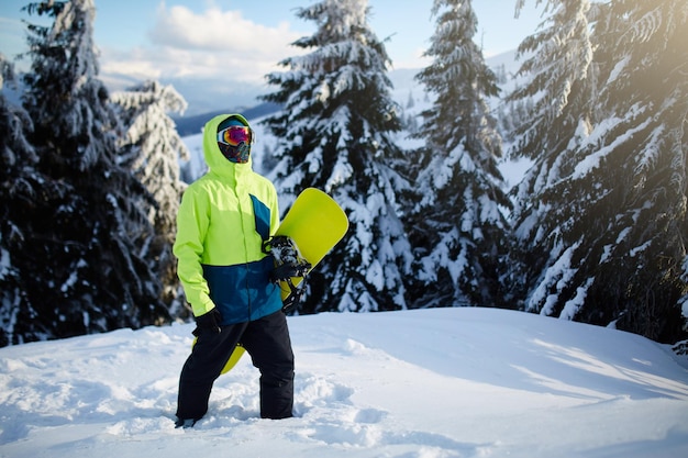 Snowboarder climbing to mountain top carrying his snowboard through forest for backcountry freeride and wearing reflective goggles colorful fashion outfit and balaclava at ski resort winter sports