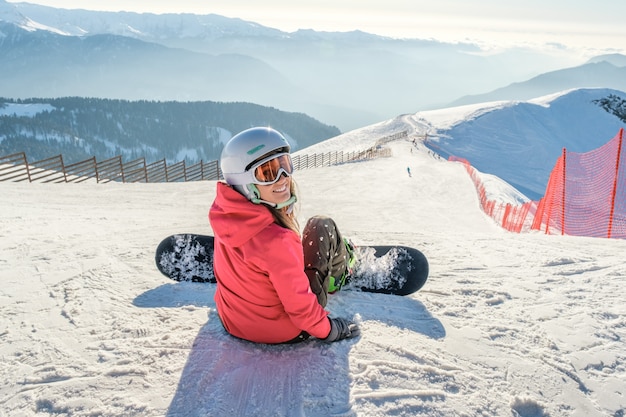 Snowboarder girl sits with board at the ski slope in mountains