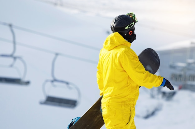 Snowboarder is walking on a snow-covered mountain. He is wearing a mask, helmet, goggles and a yellow jumpsuit