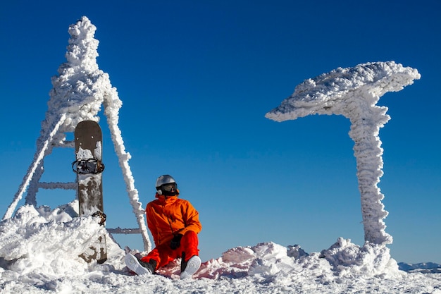 Snowboarder in a mask next to a sign covered with snow wind work in the winter mountains