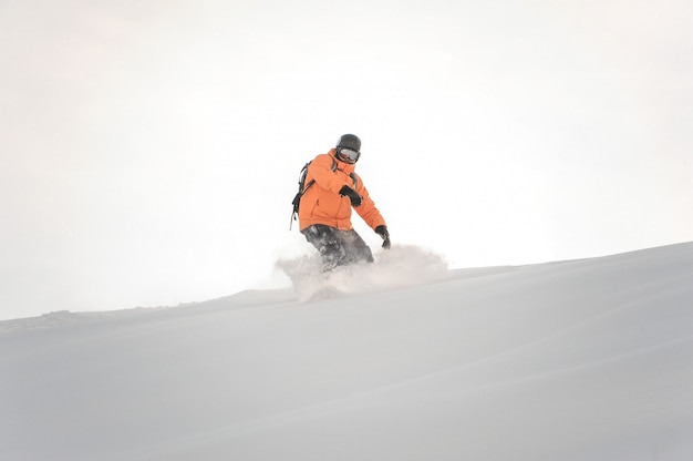 Snowboarder in orange sportswear riding down the mountain slope against the white sky