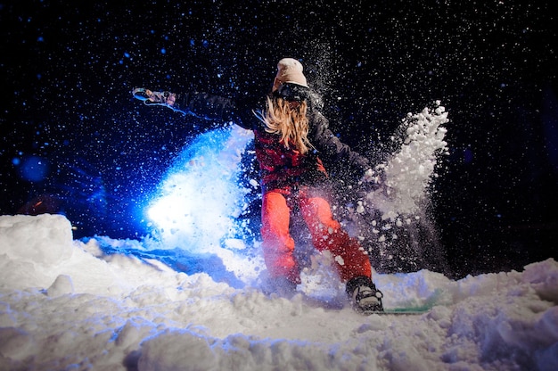 Donna dello snowboarder vestita con un abbigliamento sportivo rosso che cavalca sul pendio della montagna nella notte sotto la luce blu