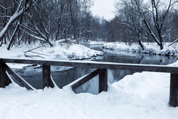 Snowcovered wooden bridge over a small river surrounded by dark bare trees