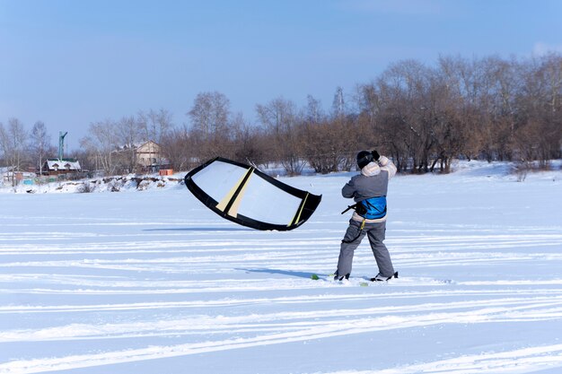 Сноукайтер ловит ветер своим воздушным змеем, стоя на льду замерзшего пруда у берега