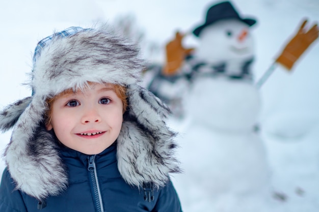 Снеговик и забавный ребенок-друг стоит в зимней шапке и шарфе с красным носом. Лепить снеговика