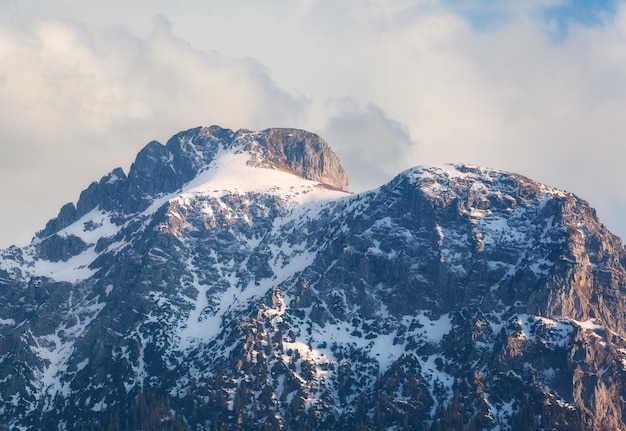 雪山は春の日没時にピークに達します。雪、森、夕方には雲と青い空に覆われた高い岩の山の風景。ドイツのアルプス。自然と旅行の背景