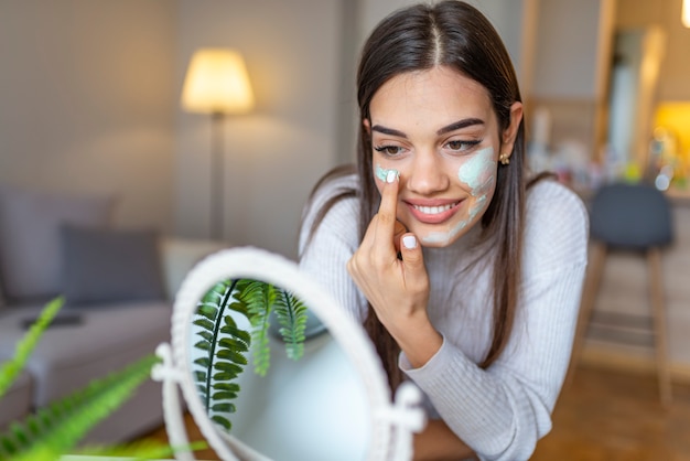 Photo so beautiful girl with beauty mask on her face looking in mirror. beautiful woman applying natural homemade facial mask at home