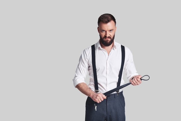 So cruel! Angry young man in formalwear carrying a belt and looking at camera while standing against grey background