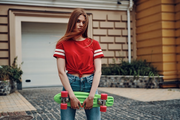 とてもかわいい！カメラを見て、屋外に立っている間スケートボードを運ぶ美しい若い女性