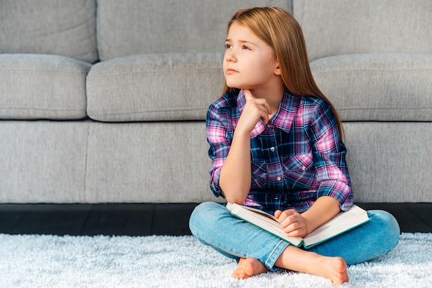 So many questions. Pensive little girl holding book and looking thoughtful while sitting on the carpet in lotus position at home