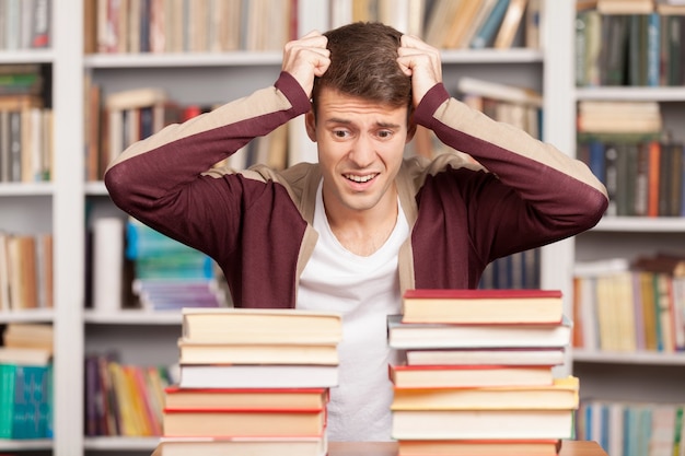 Tanto lavoro avanti! giovane stanco che si tiene la testa tra le mani e guarda le pile di libri mentre è seduto alla scrivania della biblioteca