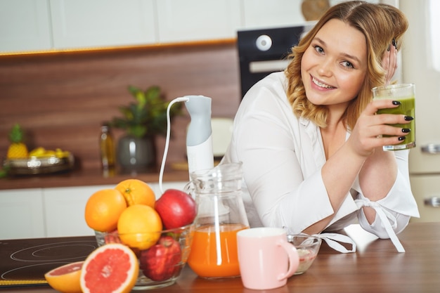 So pleased. Joyful woman leaning elbows on the table, holding glass with green smoothie