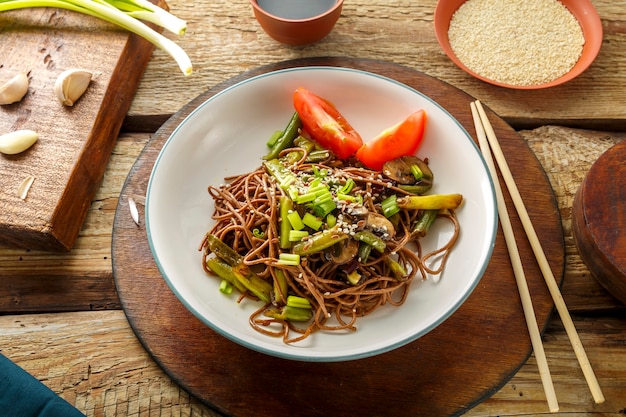 Soba with mushrooms, green beans and sesame seeds in a plate on a stand on a wooden table next to chopsticks and soy sauce and garlic on stands. Horizontal photo