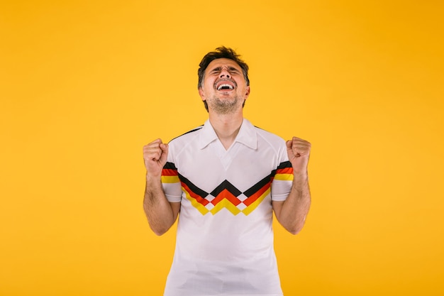 Soccer fan wearing a white t-shirt with black, red and yellow stripes, clenches his fists and yells cheering for his team.