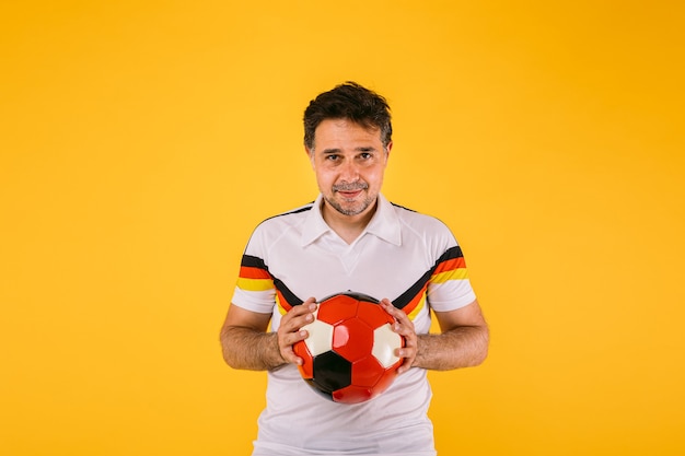 Soccer fan wearing a white t-shirt with black, red and yellow stripes, holds a ball in his hands