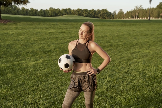 Soccer girl beautiful sporty young woman holding soccer ball\
and smiling at camera while standing