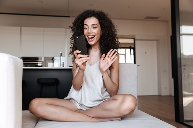 sociable adorable woman sitting with legs crossed on sofa at home, and waving hand while using mobile phone for video call