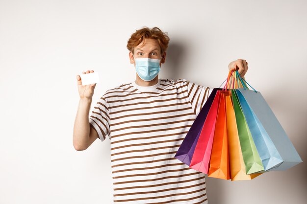 Social distancing, coronavirus concept. Young man with red hair, wearing face mask, showing shopping bags and plastic credit card, white background.