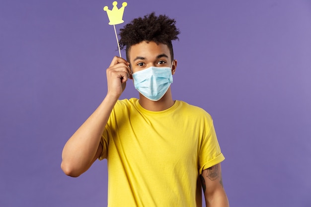 Social-distancing, coronavirus, health concept. Portrait of upbeat, happy hispanic man in medical face mask, holding small paper crown above head, staying home during quarantine, playing games