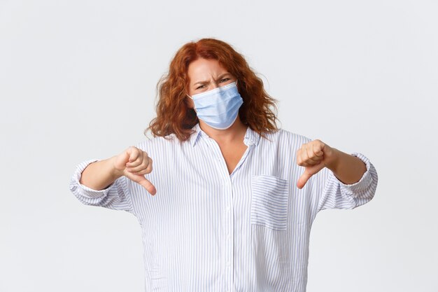  social distancing, coronavirus preventing measures and people concept. Displeased and upset middle-aged redhead woman showing thumbs-down disappointment, wearing medical mask.