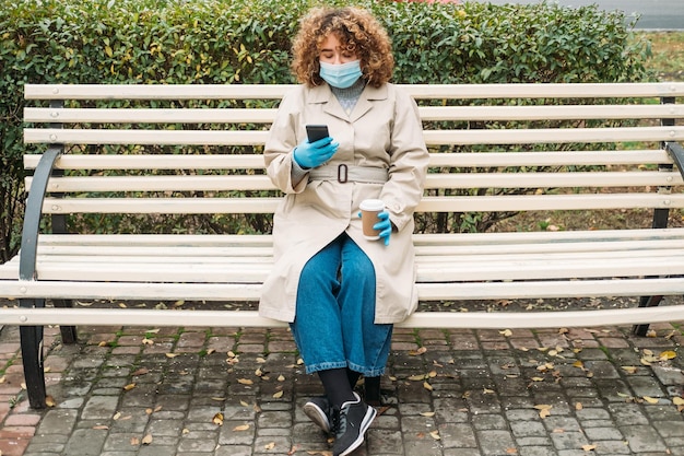 Social distancing Covid impact Pandemic loneliness Young overweight woman with curly hair in protective face mask phone texting on bench in city street