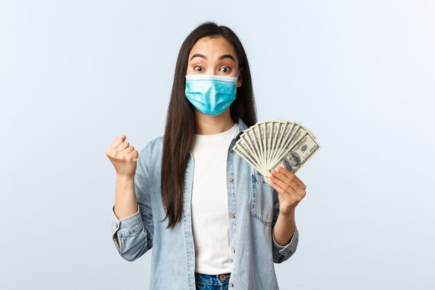 Social distancing lifestyle, covid-19 pandemic business and employement concept. Excited happy asian woman winning lottery, wear medical mask and rejoicing, show money, holding cash