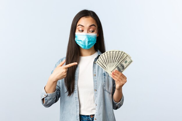 Social distancing lifestyle, covid-19 pandemic business and employement concept. Excited pleased asian girl in medical mask pointing finger at cash, found big money