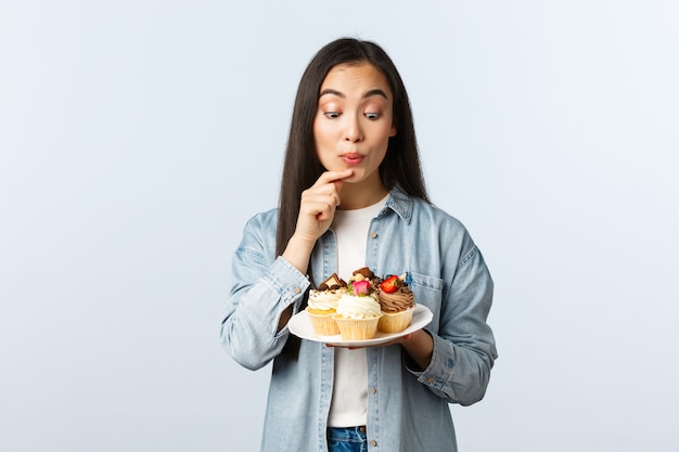 Social distancing lifestyle, covid-19 pandemic, celebrating holidays during coronavirus concept. Delighted and temting cute asian girl staring with desire at plate of cupcakes