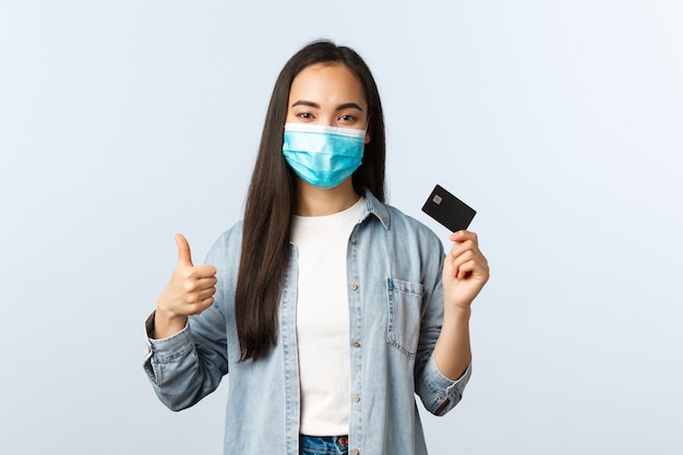 Social distancing lifestyle, covid-19 pandemic and contactless shopping concept. Satisfied asian female in medical mask show thumbs-up gesture and promote credit card features, bankc account