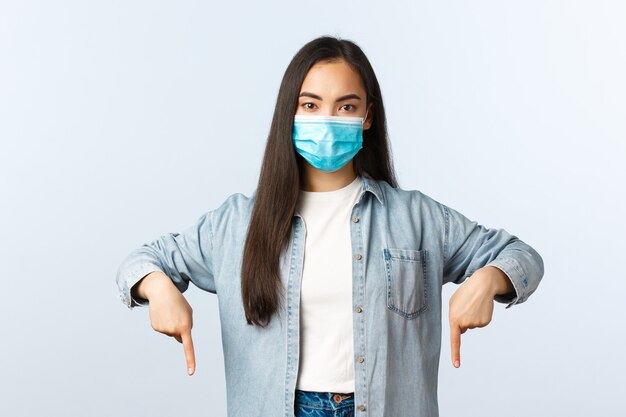 Social distancing lifestyle, covid-19 pandemic everyday life and leisure concept. Confident and determined young asian woman in medical mask pointing fingers down in casual outfit.