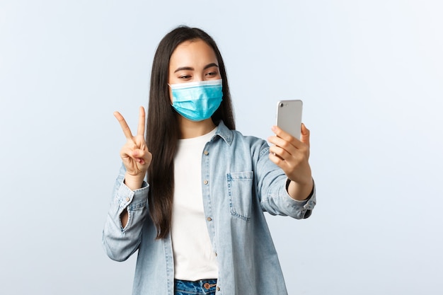 사회적 거리를 두는 생활 방식, covid-19 전염병 및 사람들의 감정 개념. 의료용 마스크를 쓴 친근하고 쾌활한 아시아 여성이 소셜 미디어를 위해 셀카를 찍고 스마트폰 카메라에서 평화 서명을 합니다