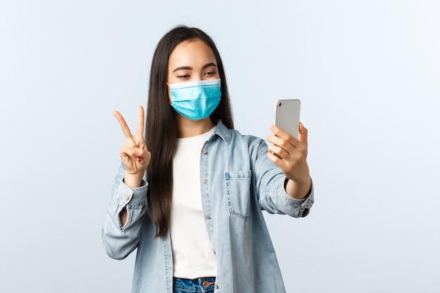 Stile di vita di distanza sociale, pandemia di covid-19 e concetto di emozioni delle persone. amichevole donna asiatica allegra in maschera medica che prende selfie per i social media, fa il segno della pace alla fotocamera dello smartphone