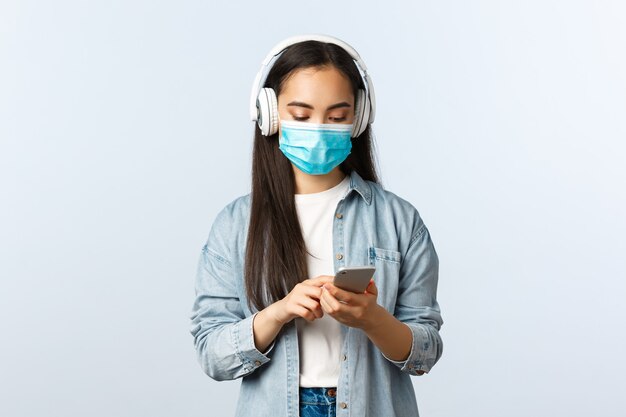 Social distancing lifestyle, covid-19 pandemic and self-isolation leisure concept. Modern cute asian girl in medical mask, listening music wireless headphones, using mobile phone app