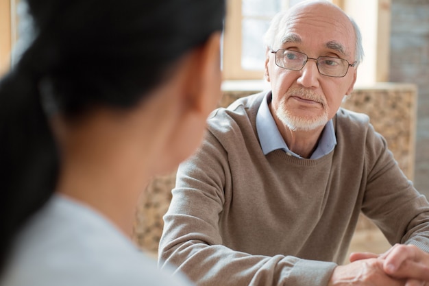 Photo social interaction. handsome positive senior man wearing glasses while listening to volunteer and posing on blurred background