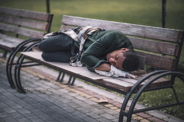 Social issues. Dark-skinned young adult man with closed eyes lying on bench in park during day