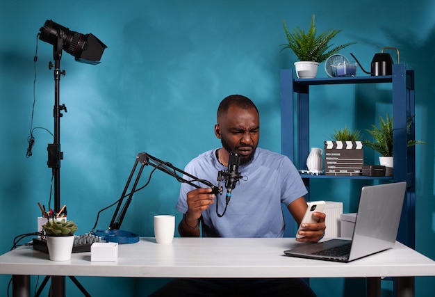 Social media influencer frowning after reading upsetting message on smartphone during live podcast. Vlogger sitting at desk in vlogging studio recieving disturbing question from audience.