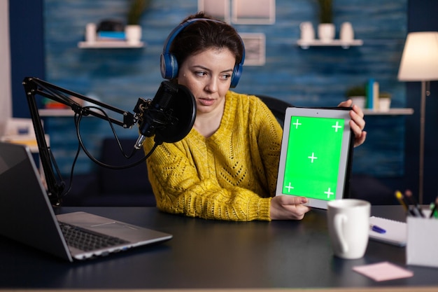 Social media influencer recording online video for\
entertainment vlog holding mock up green screen chroma key tablet\
with isolated display in living room. vlogger woman using\
podcasting equipment