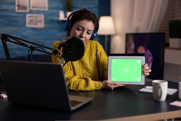 Social media influencer talking with fans into microphone showing mock up green screen chroma key tablet with isolated display at camera during online livestream. Vlogger recording on-air show