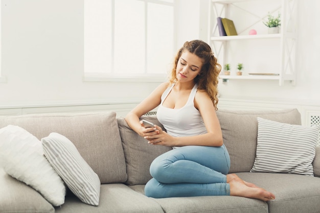 Sociale netwerken. Jonge roodharige vrouw berichten thuis op smartphone, comfortabel zittend op een beige bank, kopieer ruimte