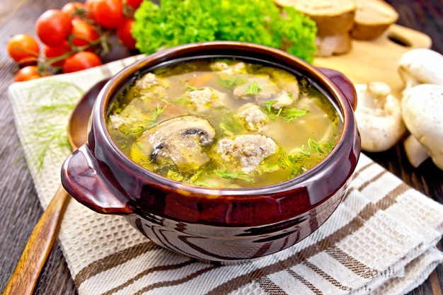 Foto soep met gehaktballen, noedels en champignon in een kleikom op een servet, peterselie, tomaten, champignons en brood op houten plankenachtergrond